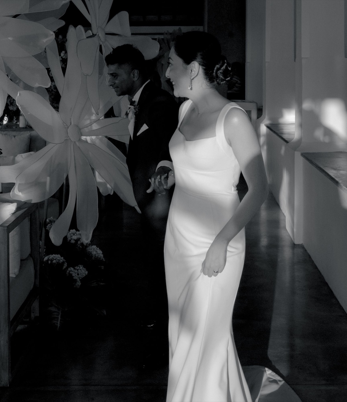 A bride and groom walking hand in hand past large white paper flowers, in black and white