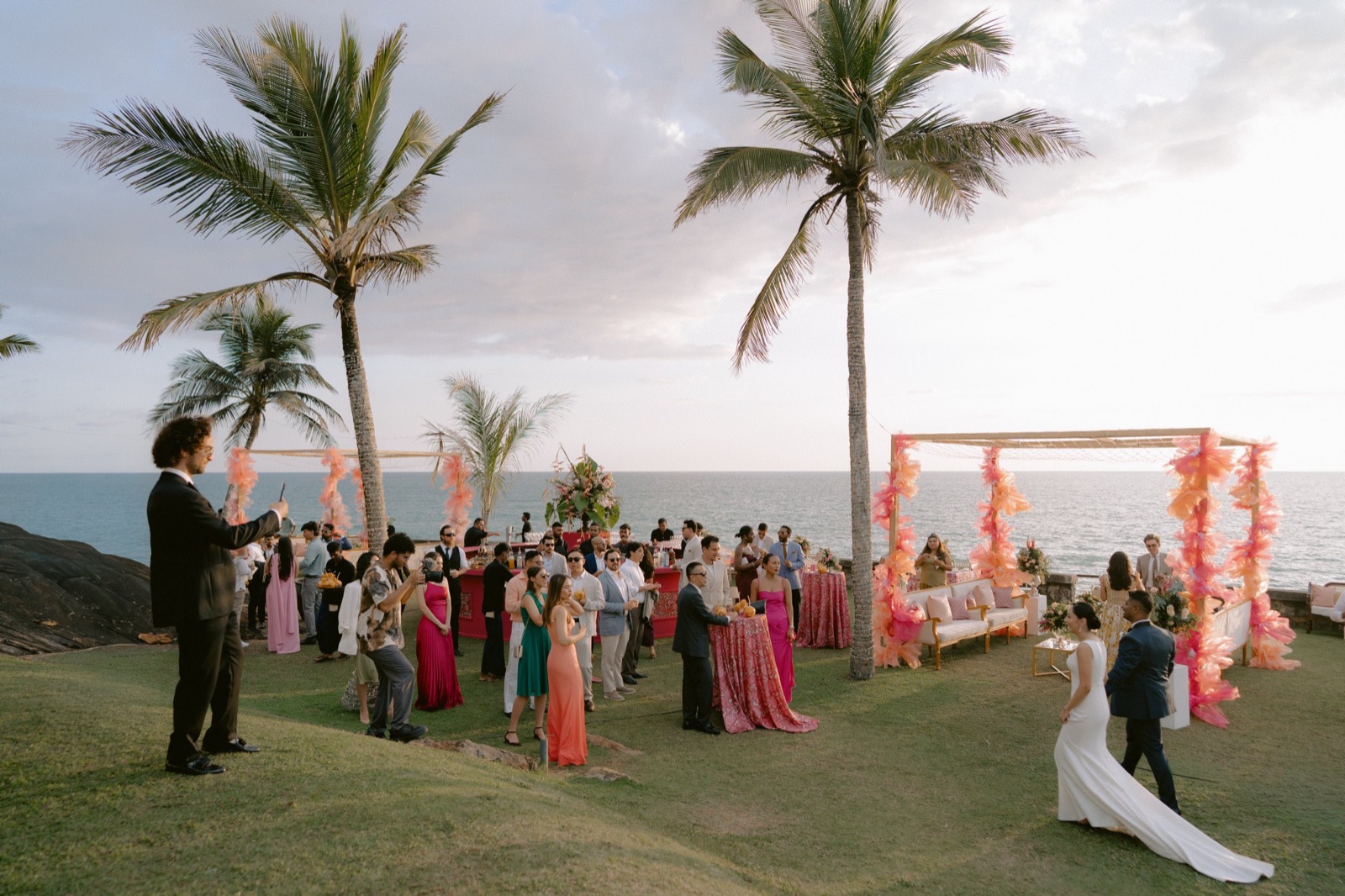 Guests on the clifftop lawn above the sea at sunset, the bride and groom walking in