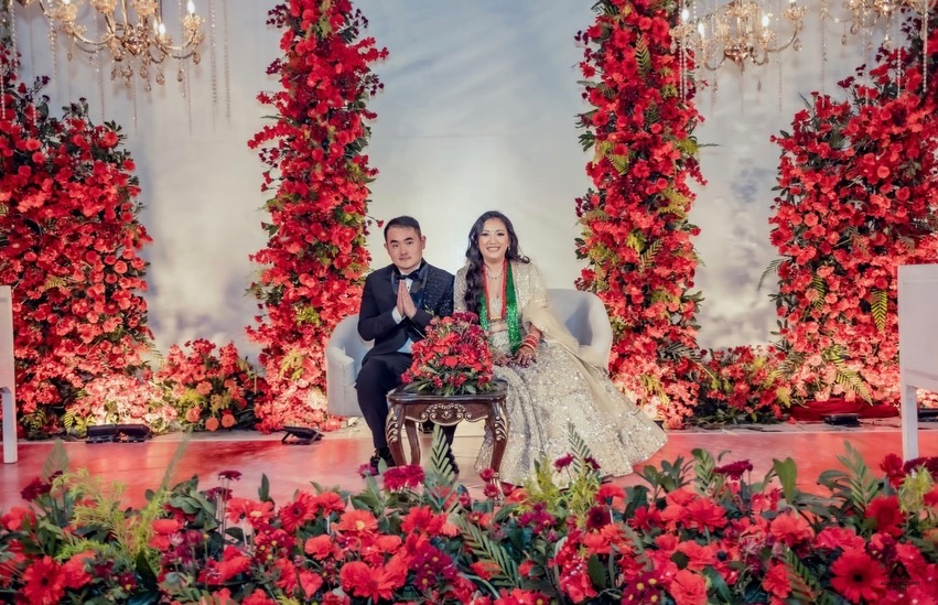 Kusum and her husband seated on a stage between walls of red flowers