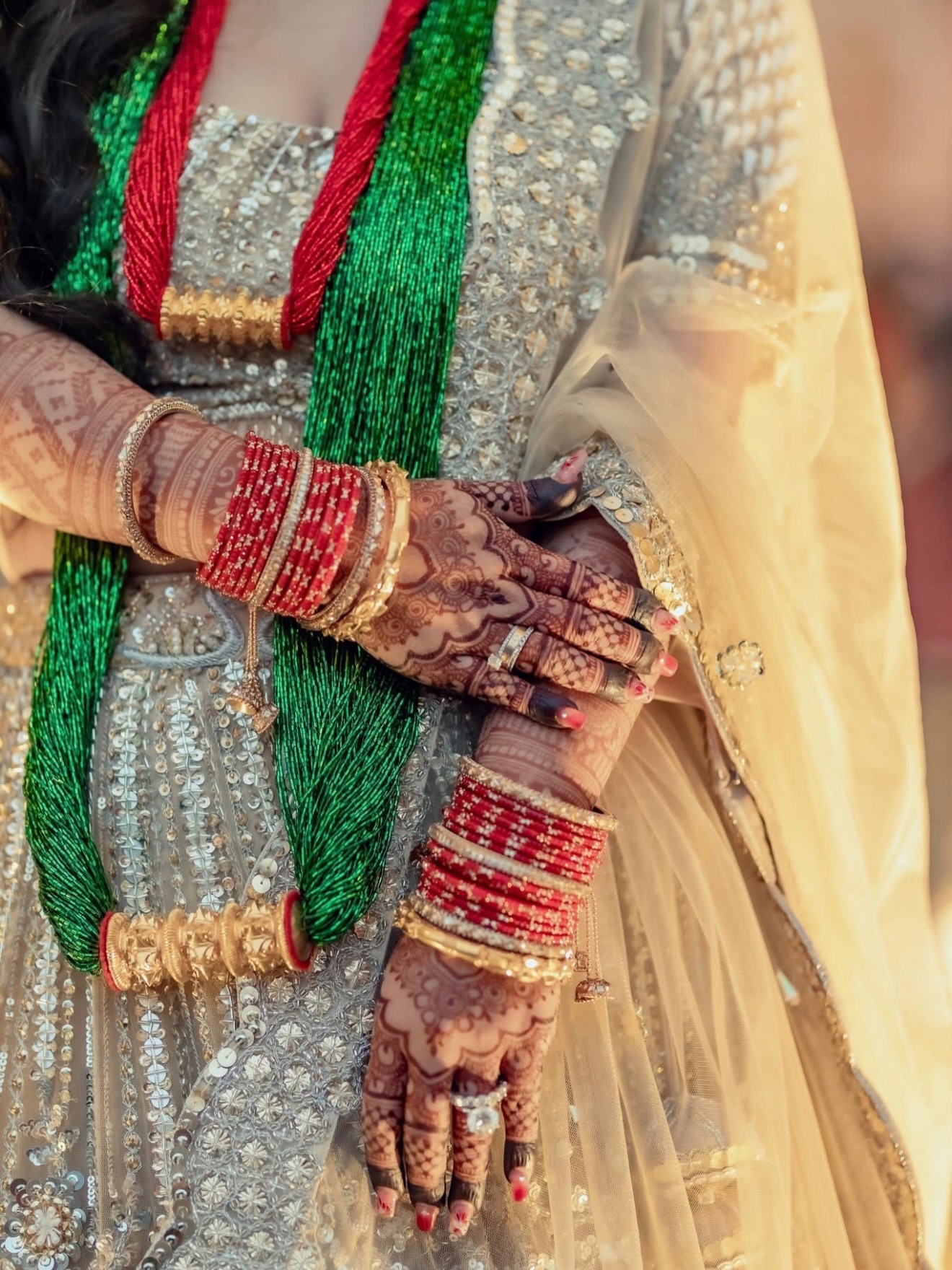 Hennaed hands with red bangles over a green bead necklace and a gold lehenga