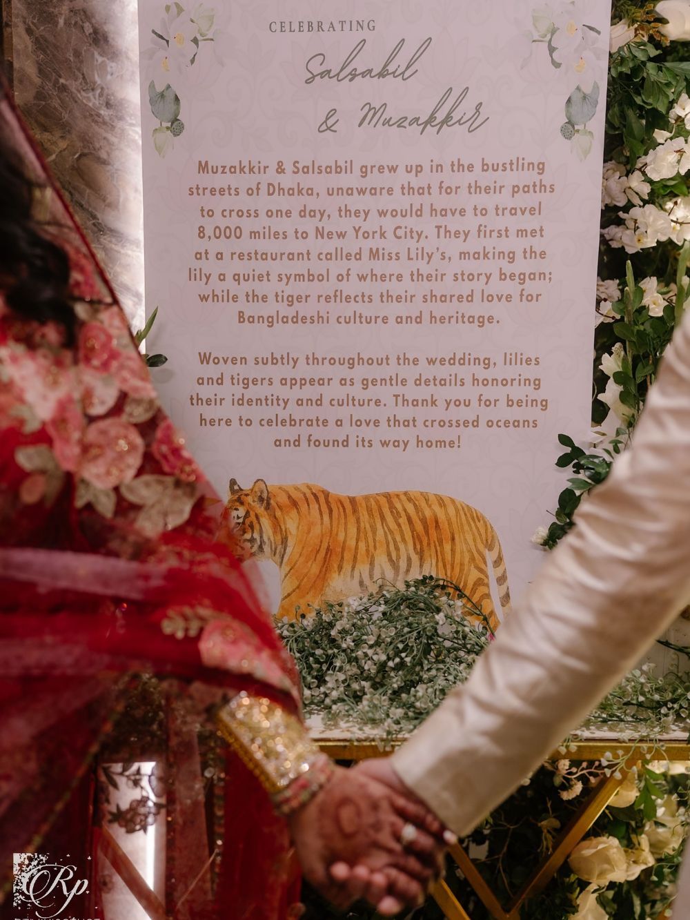 The couple holding hands beside a welcome sign painted with lilies and a tiger