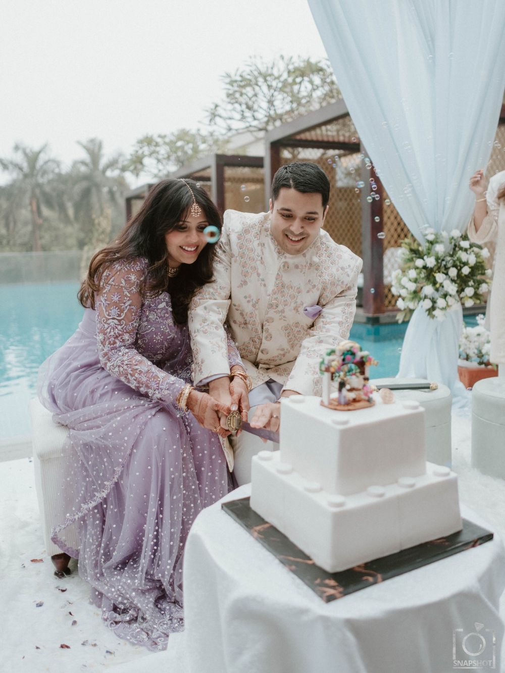 Salsabil in lilac and her husband cutting a white cake by a pool