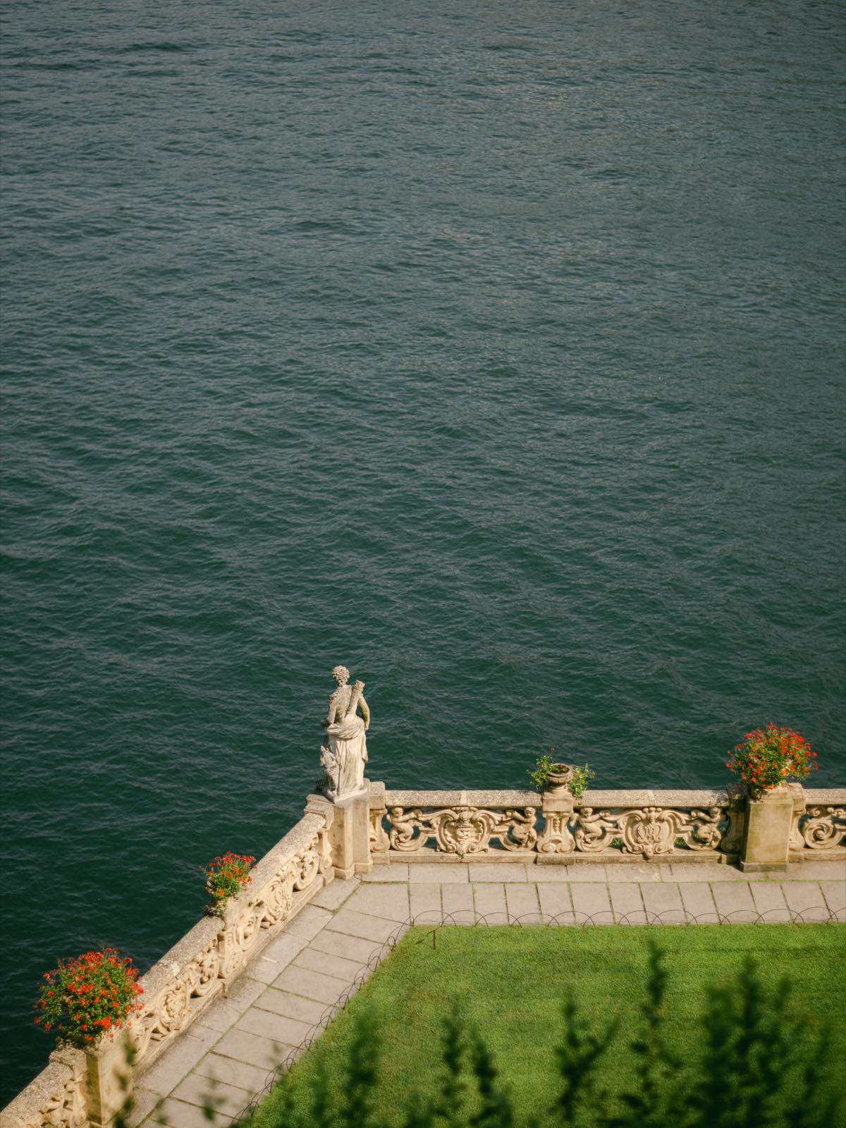A stone terrace with a statue and red geraniums above the lake